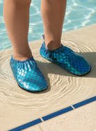 Blue mermaid scale-patterned Ivy shoes worn by a person standing on sand near water.